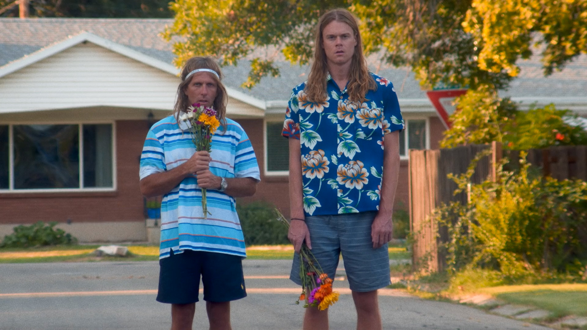 Jer and MP standing next to each other on a road in a neighbourhood. Jer is standing holding a firm grip on a bouquet of flowers close to his body, while MP is holding a similar bouquet in one hand as if he's about to let go of the flowers to the ground. They are both looking ahead of themselves with disappointed faces. 