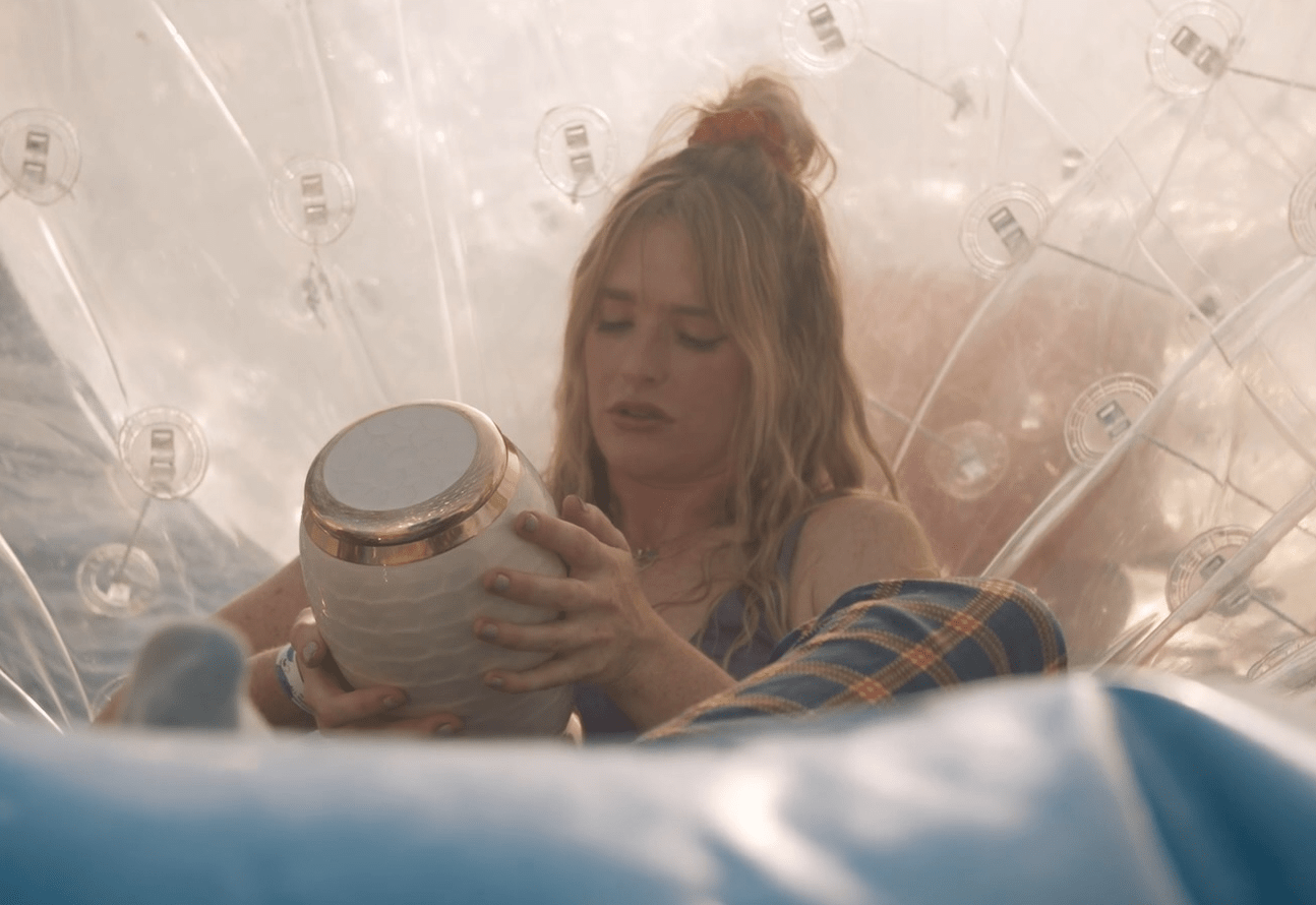 A woman (Kate Jean Hollowell) sitting inside a plastic bubble at the funfair. She is holding and talking to her parents, whose ashes are in the urn she is holding up towards her face. 