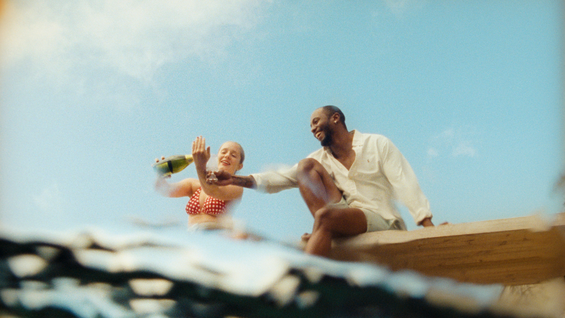 A low angle shot from just beneath the surface of the water, as the camera looks up towards a woman and man sitting together laughing and drinking alcohol.