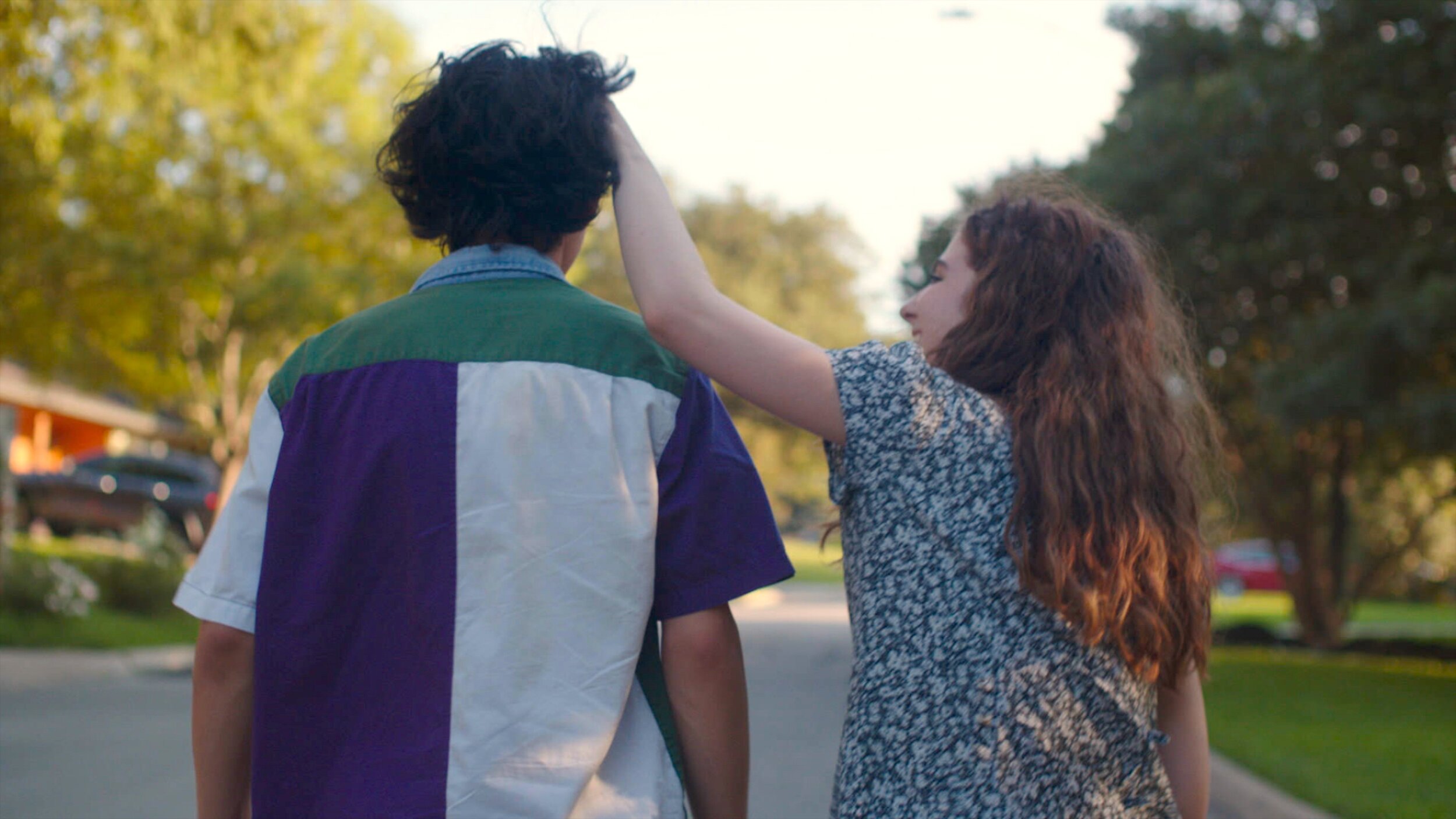A boy and girl shown from behind as they are walking down a neighbourhood street. She is looking up towards him, laughing, whilst touching the boy's dark hair. 