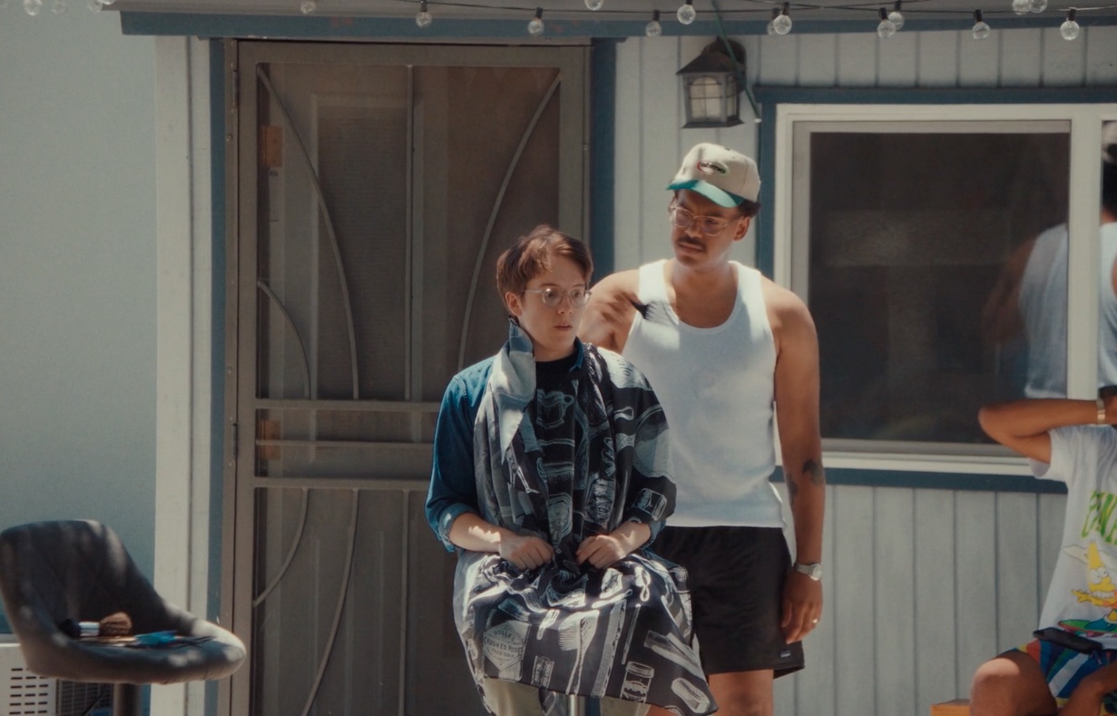 A still from the story about a backyard barber. On a stool a nervous-looking young man is sitting whilst a man is standing behind him combing through his hair. 