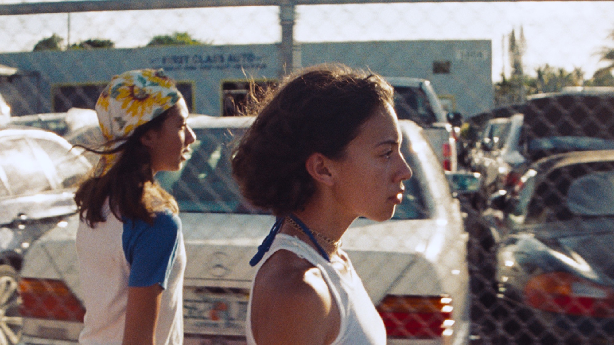 Adrian and her younger sister seen in profile walking next to a fence with rows of cars behind it. Adrian looks focused, as if she is looking towards a goal in the distance. Her younger sister has a more casual body language, underlining how she is just following along but not interested in the teenage group they are following.