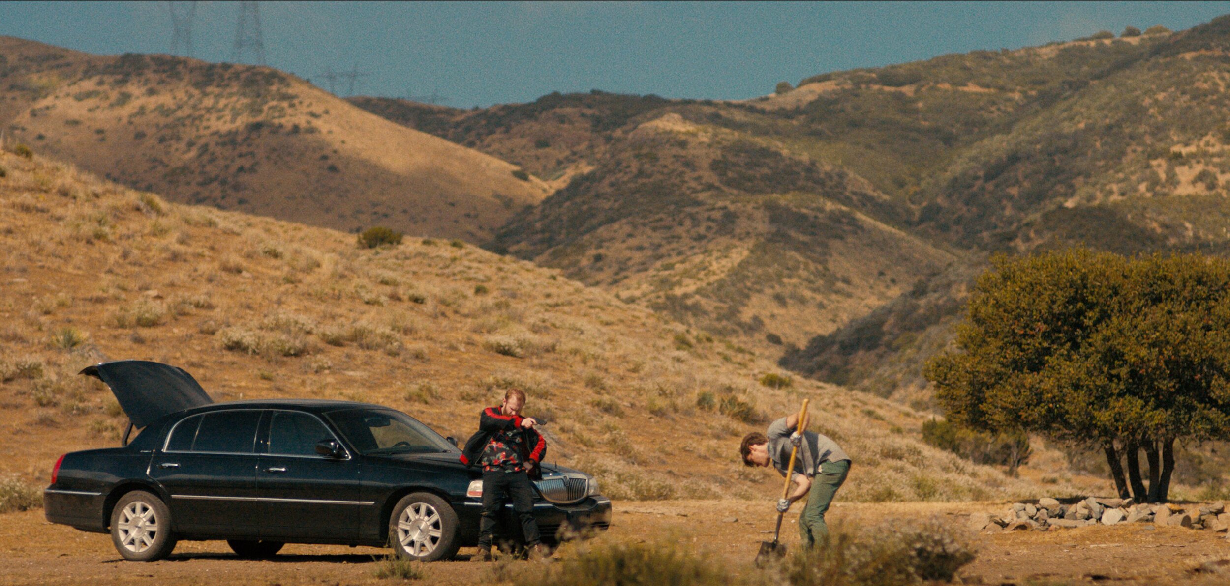 Still showing the two main characters from a distance. The hitman is leaning against his black car (which has the trunk open) with a gun in his hand aimed towards the other man. The other man is trying to dig. The environment is scarce and dry with overwhelming hues of brown and beige and just a little hint of green. 