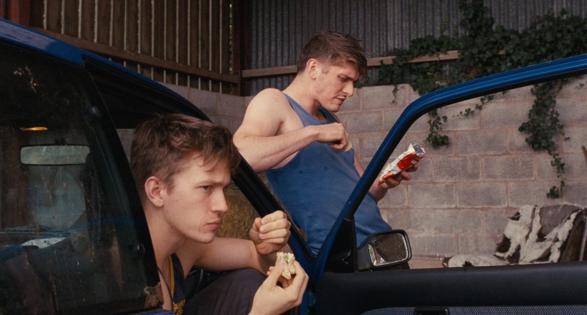 Rhys and Freddie taking a break from work. Rhys is sitting in a car with the door open, eating a sandwich and looking very concentrated ahead. He looks tense. Freddie is leaning against the hood of the car, reading on the back of a packet of crisps. He looks concentrated as if the text is too small. 