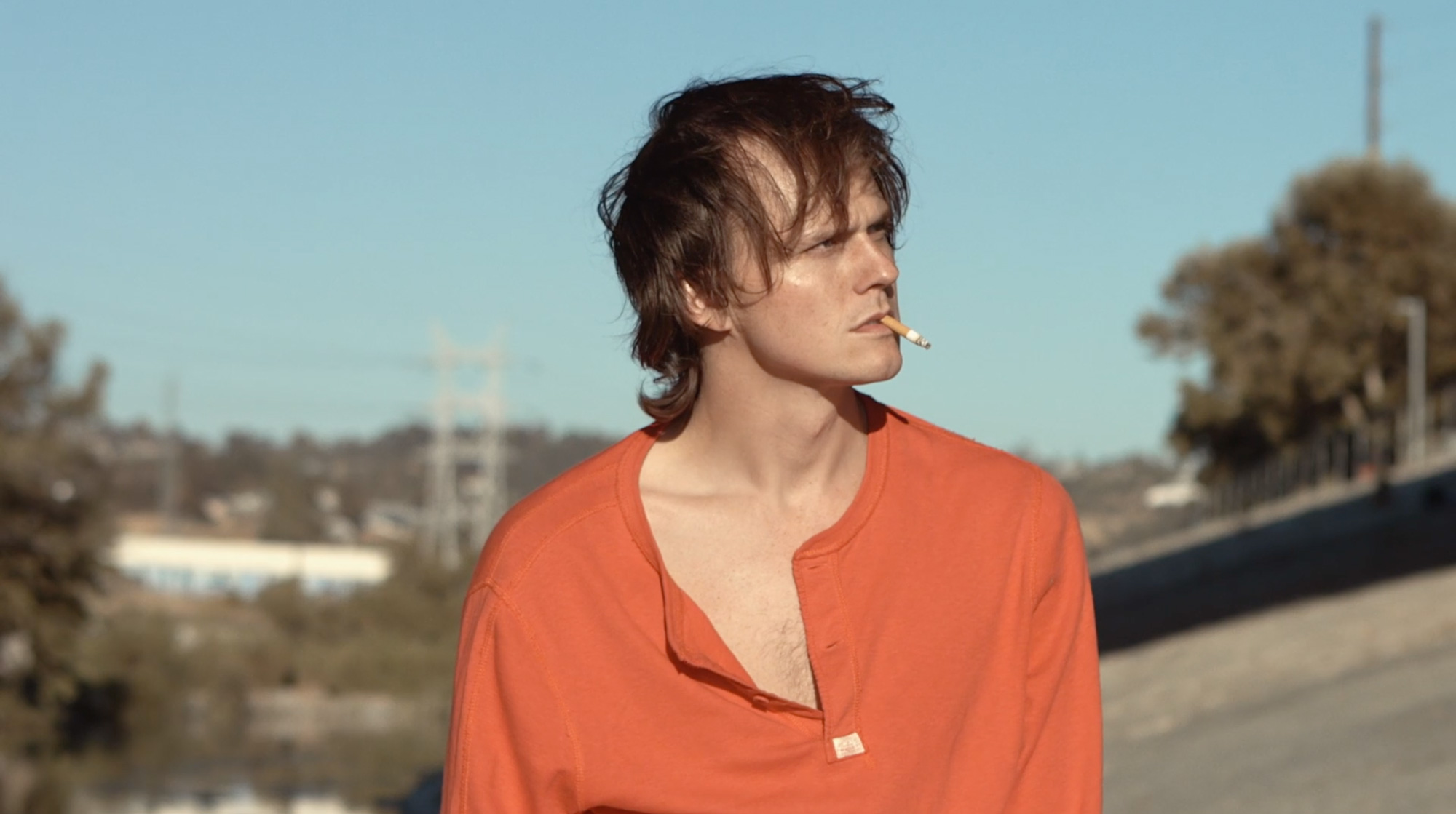 Whit in focus as he is walking through LA during daytime looking annoyed. He is smoking a cigarette, it casually hanging out of his mouth, his brown hair sits messy on top of his head and his light red/orange shirt is unbuttoned to expose some of his chest. 