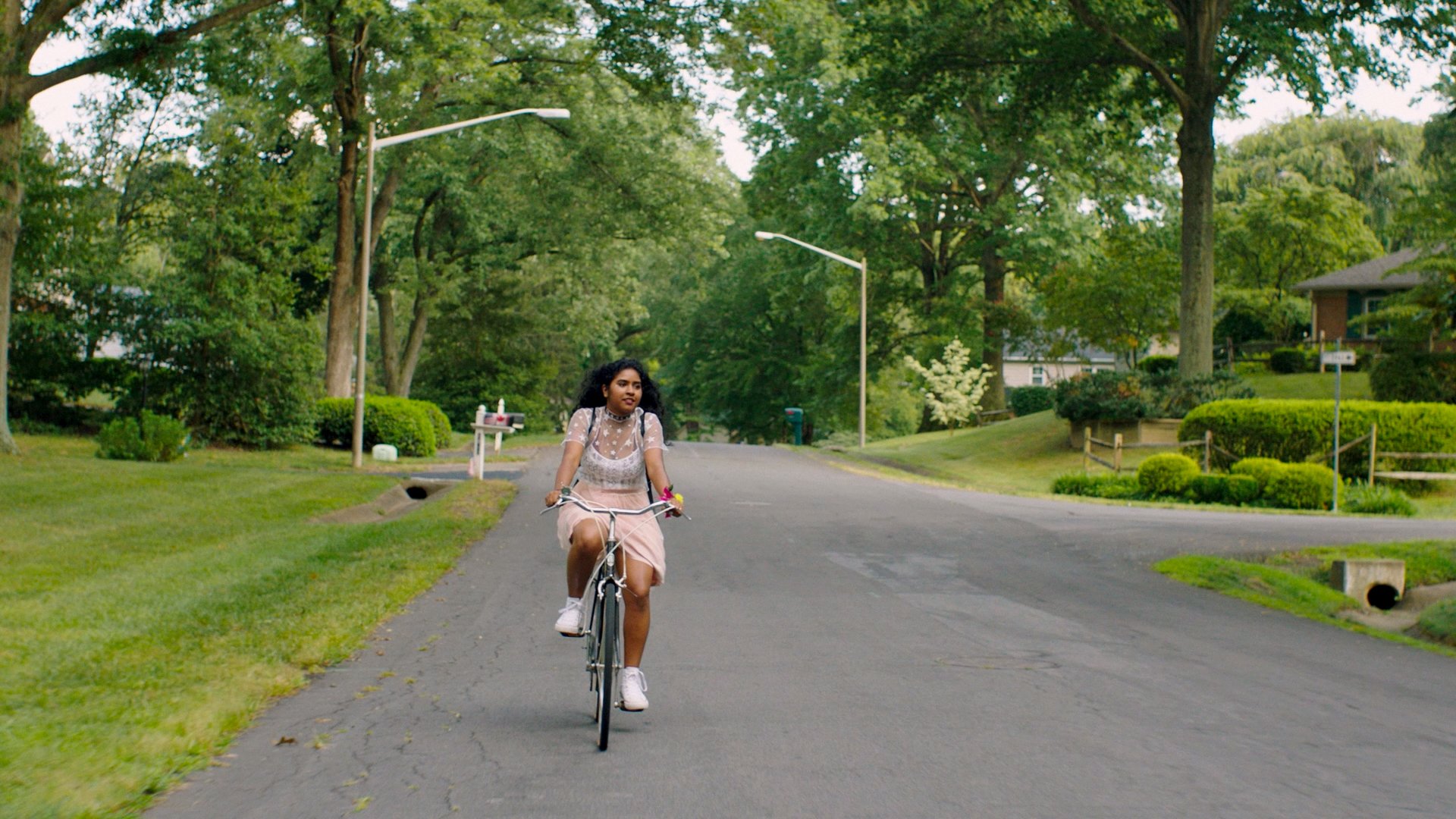Lekha riding her bike after sneaking out of her house. She is riding it through a pretty neighbourhood with lots of greenery and trees following the road she is following. 