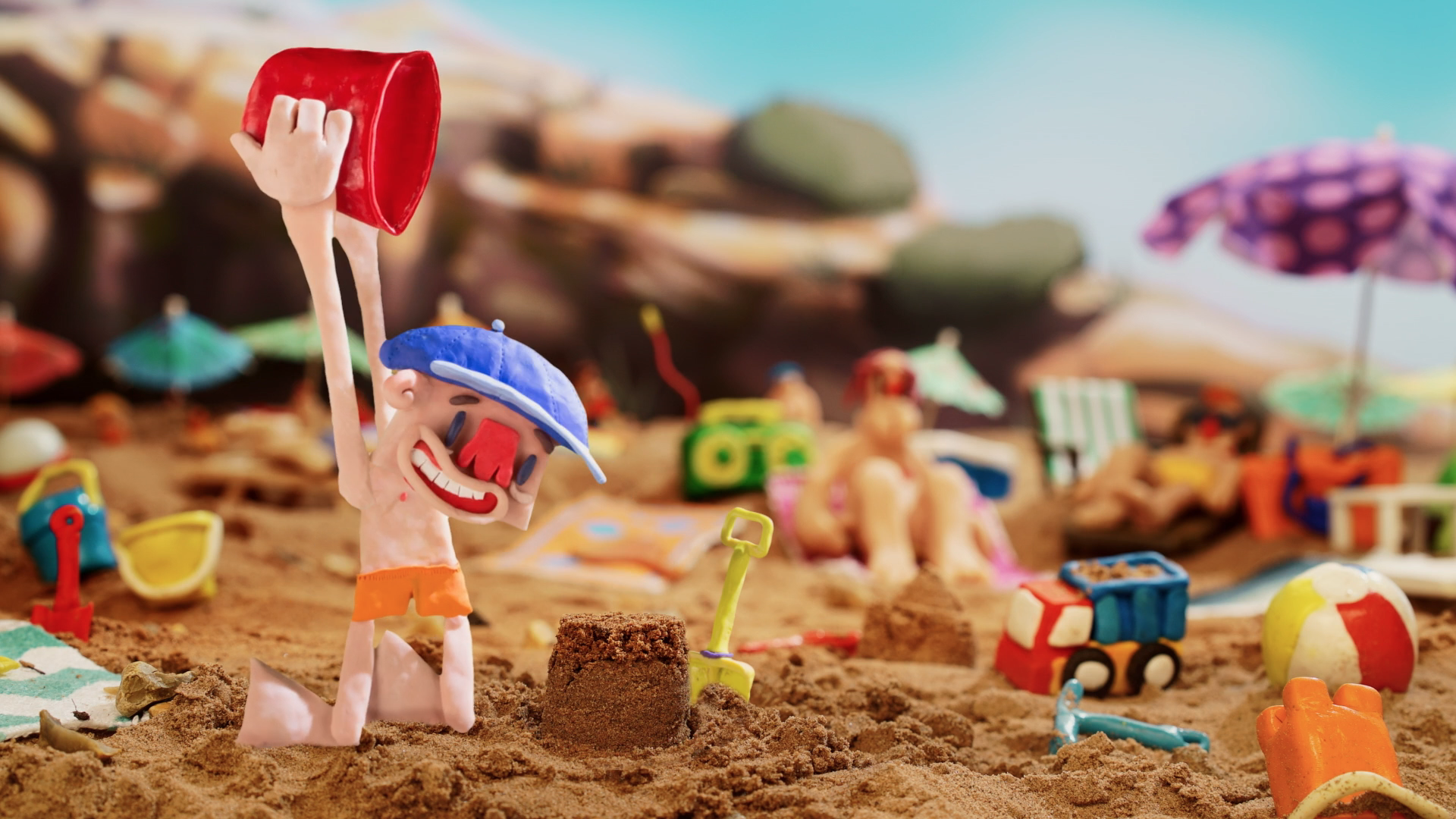 A little boy is happy as he is building a sandcastle on the beach. He is wearing orange swim trunks and a blue cap. The bucket he is holding up over his head is red and his shovel is yellow. He is in focus, behind him are the outlines of various other objects and people seen, including people sunbathing in their chairs, other toys and a radio. 