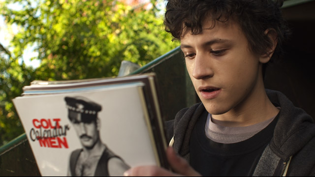 Jack shown holding up a bunch of magazines with gay porn. He looks in awe with his mouth open. Curious. On the back of the last magazine of the bunch the still shows a man dressed in a black cap and black vest and the text "Colt Calendar Men" beside him. 