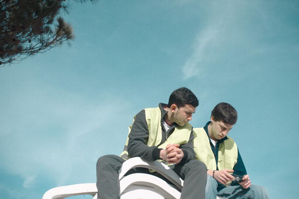 Low angel of Marc Antoine and Michael shown sitting on top of a structure as they are working. They are both wearing neon vests. The sky is blue behind them. They are looking at a video on a mobile phone. 