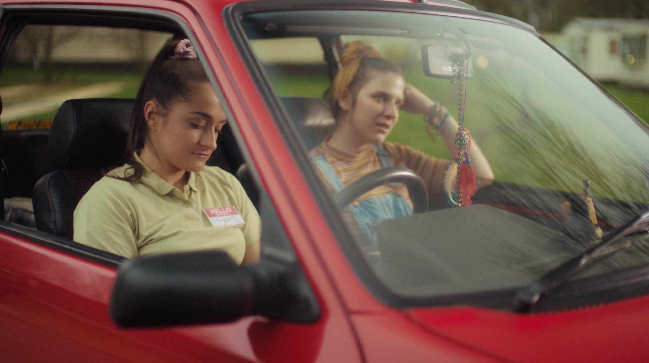 Vicky and Stacey sitting inside the car. Stacey looks down at her hands in her lap while Vicky looks ahead with a more frustrated facial expression. The details shown of the car is red (exterior) and black (interior). 