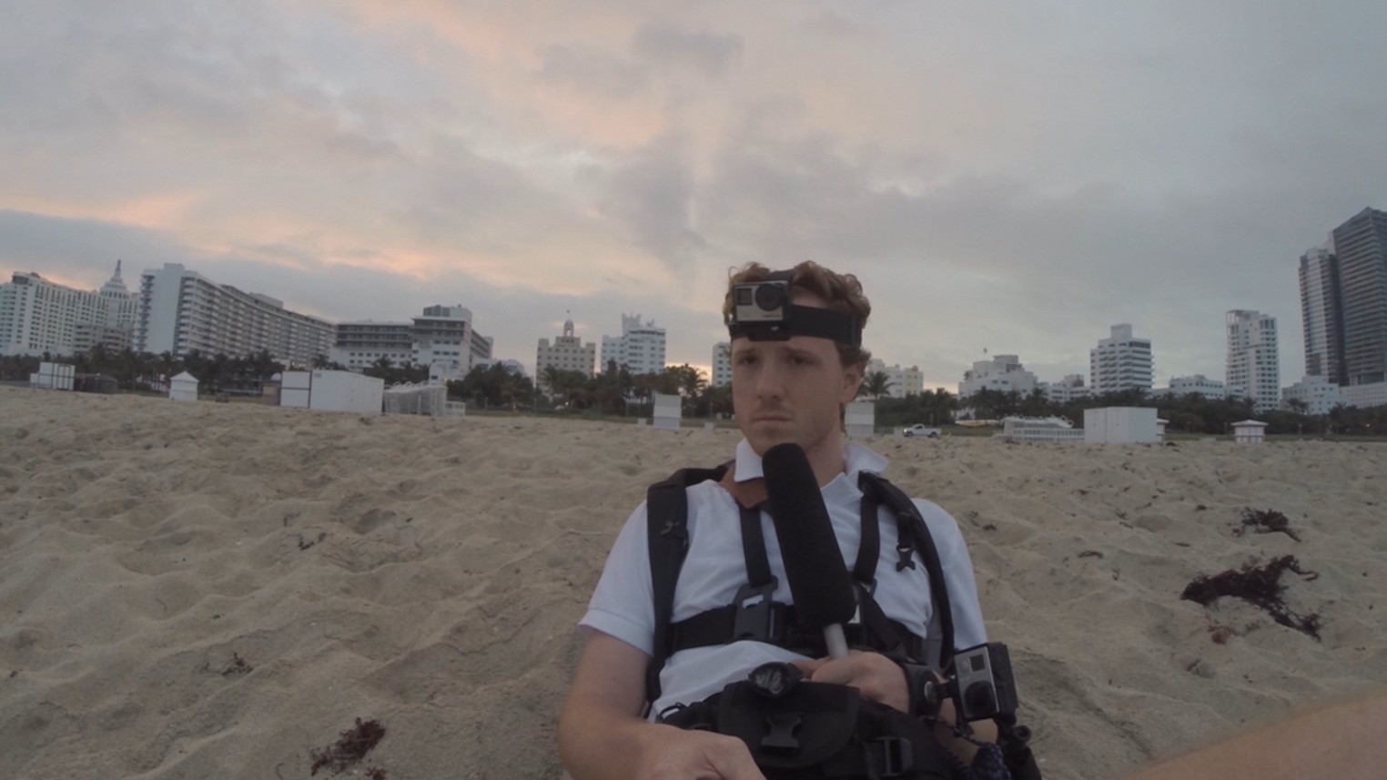 Dylan shown sitting on a beach, in the sand,  with all of his gear on (GoPros and a microphone in hand). He looks sad and disappointed, but primarily lonely as he is the only visible person on this beach. In the distance, large and tall buildings are seen. 