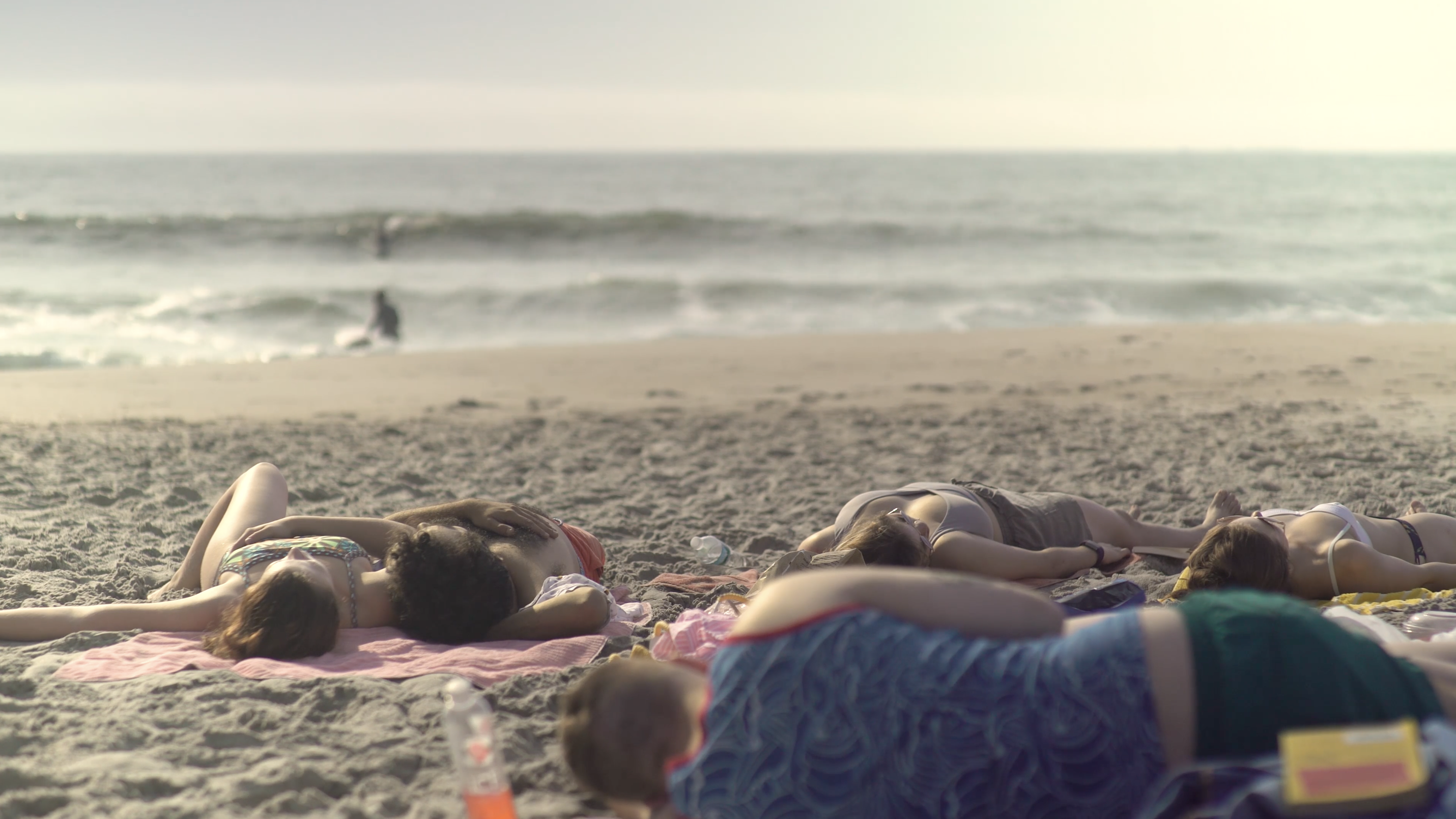 Couples and individuals shown sleeping on beach towels on the sand. They are all in various positions. It looks like they are in a haze. In the distance waves from the ocean are shown, with some people exploring them. 