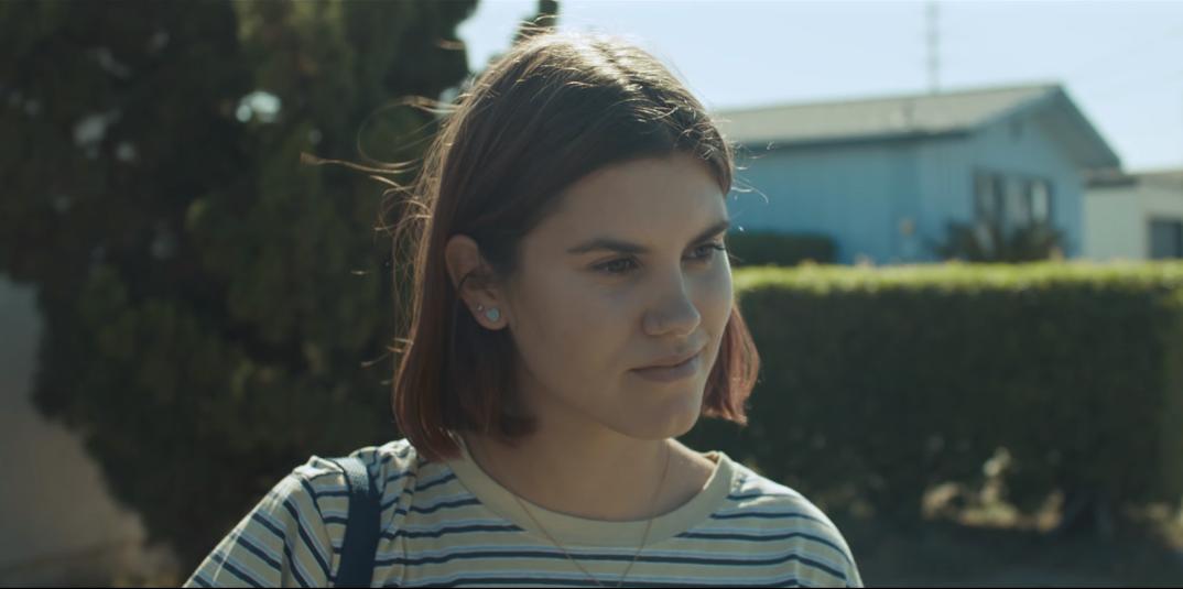 Charlotte shown during her first meeting with Tommy. She is looking at him in his car—not pictured—with a slight smile on her face. She is walking in a neighbourhood, as a row of houses are shown behind her. Behind her. arealso bushes and trees of greenery. She is wearing a striped t-shirt and her dark hair with coloured  ends are slightly above her shoulders. 