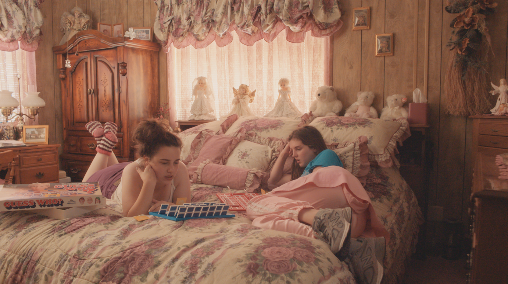 Lucy and Julie seen in a bedroom playing Guess Who?. The room is overwhelmingly pink. The curtains are white lace with pink and flowery details. The bed is covered in pillows with flowers and frills, as is the bedsheet. Behind the bed there is a shelf with decorative angels, dolls and teddy bears in white. It looks like an old home, owned by someone who has collected many things over the years.  