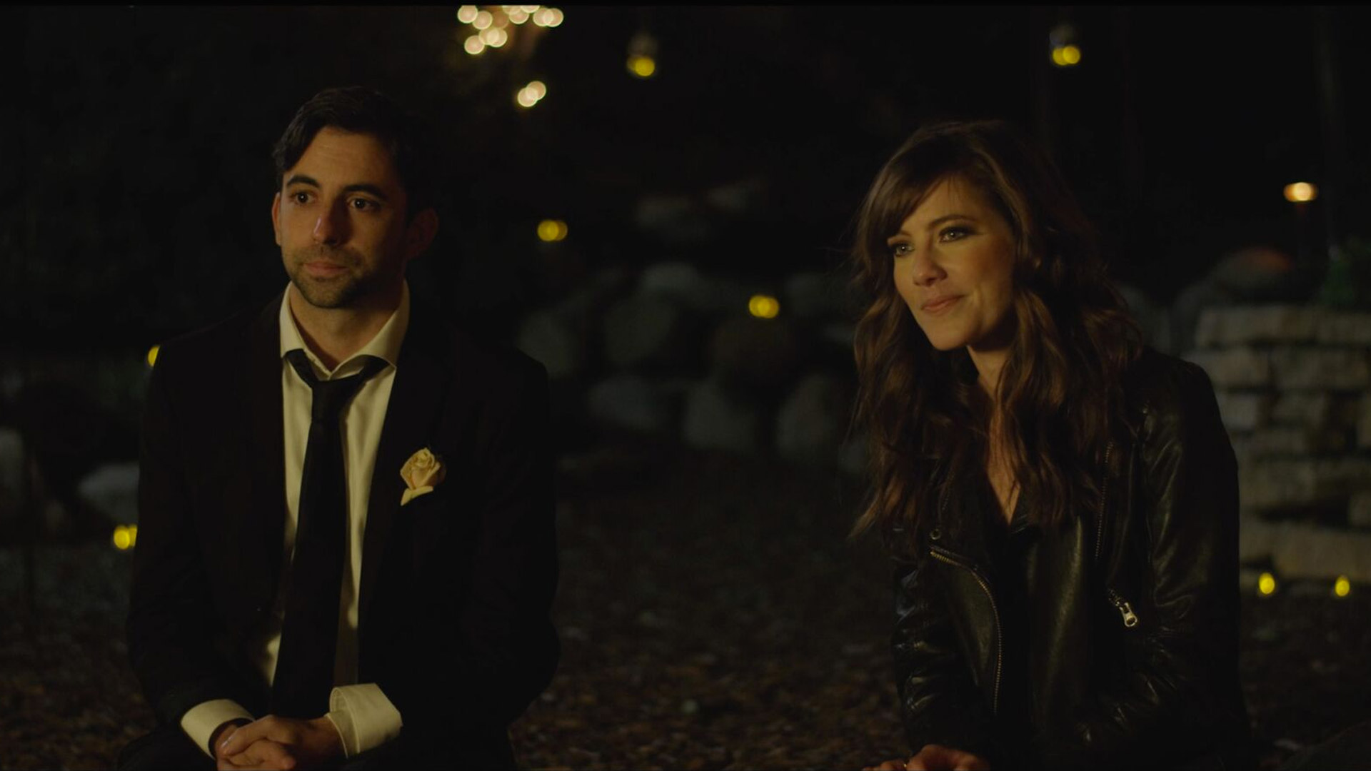 The leads sitting next to each other during nighttime looking towards the wedding reception happening in front of them. The man looks a bit collected and nervous whilst the woman has a slight smile on her face. Behind them are a couple of small light sources (candles, string lights) lighting up the still. 