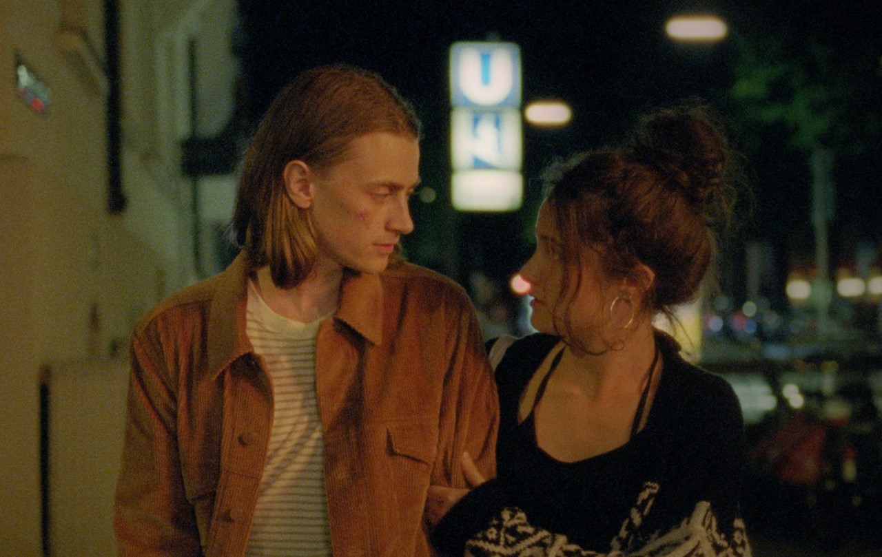 Laia and Emil seen walking down a road during the evening. Neon signs are shown out of focus behind them. They are looking at each other and Laia is holding onto Emil's arm. They look serious and contemplative. 