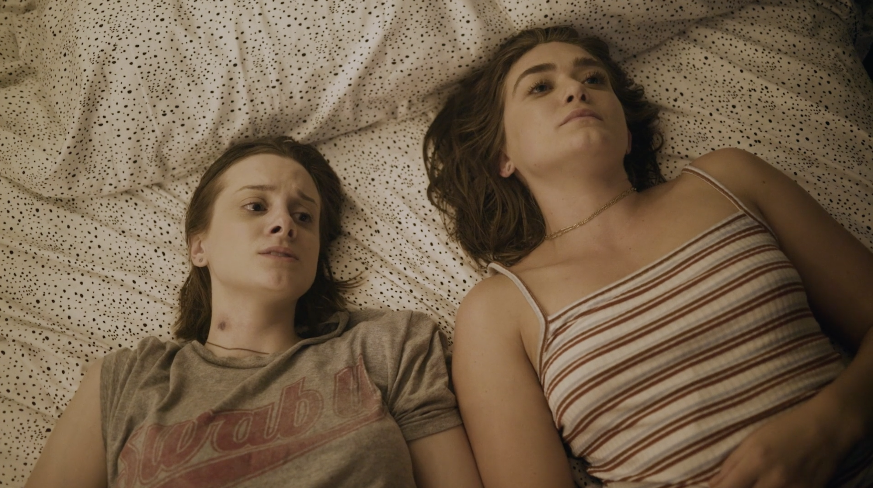 Alice and Jill shown in bed. Alice looks concerned, worried even, while Jill looks much more focused as she is looking up towards the ceiling. The bedsheet they are on top of is white with small black specks. 