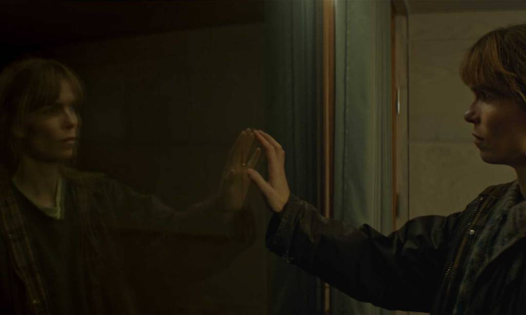 A woman looks at what appears to be her reflection in a pane of glass, resting her hand on it.