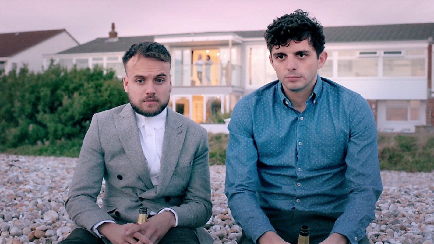 James and Mike seen sitting next to each other on the stoney beach in front of the rented house. They are dressed up but look miserably ahead with a beer bottle each in hand. Slouchy body language. In the distance, their partners are seen standing on the balcony of the house. 
