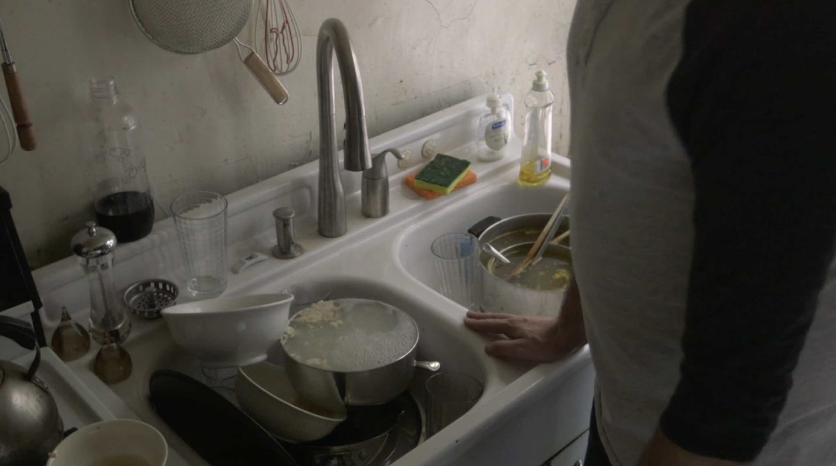 Still showing a sink overflowing with dishes. There's glasses, pots and pans, some of which have water inside of them which reveals what they contained prior to ending up in the sink (noodles etc). It looks messy as objects have continued to be placed in the sink even though there's no space. 