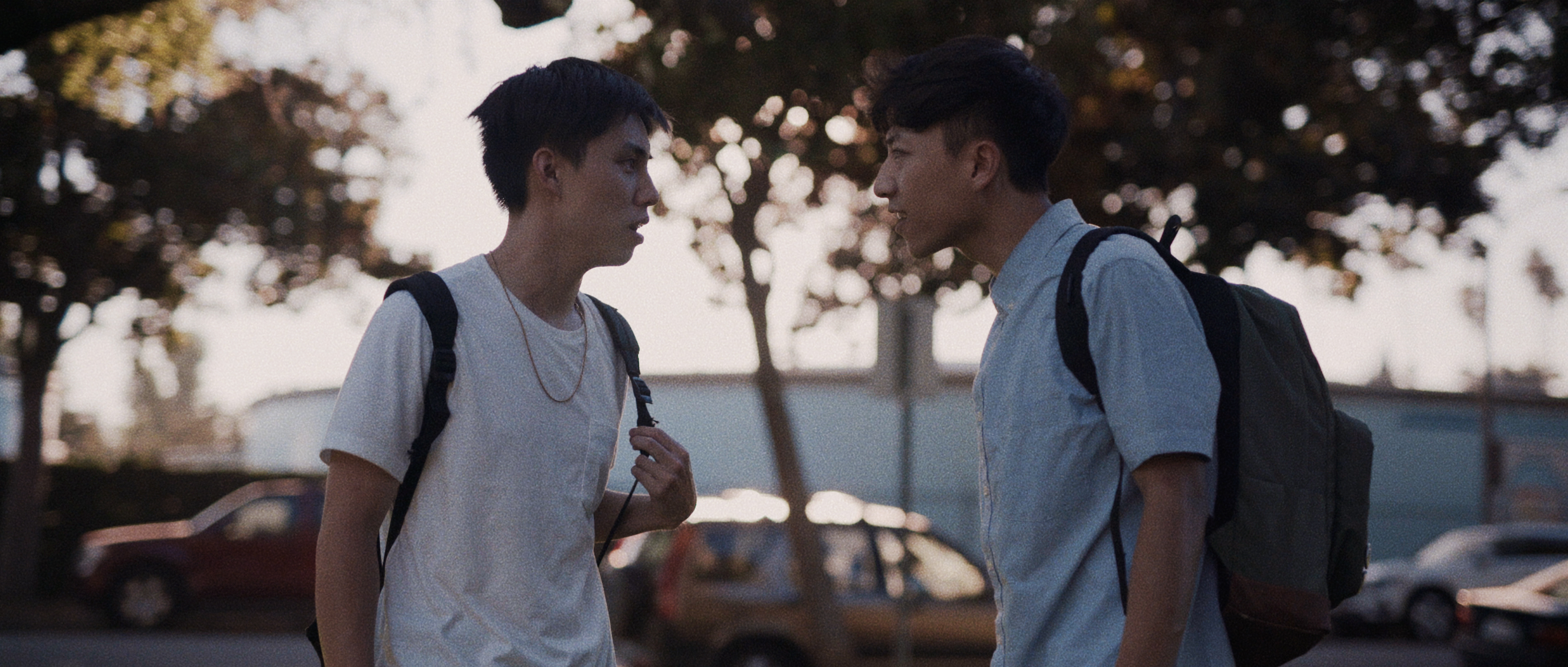 Ryan and Kev arguing on a street. They are standing opposite each other, seen in profile in this still, as their body language is slightly hostile and argumentative. They are both wearing backpacks. 
