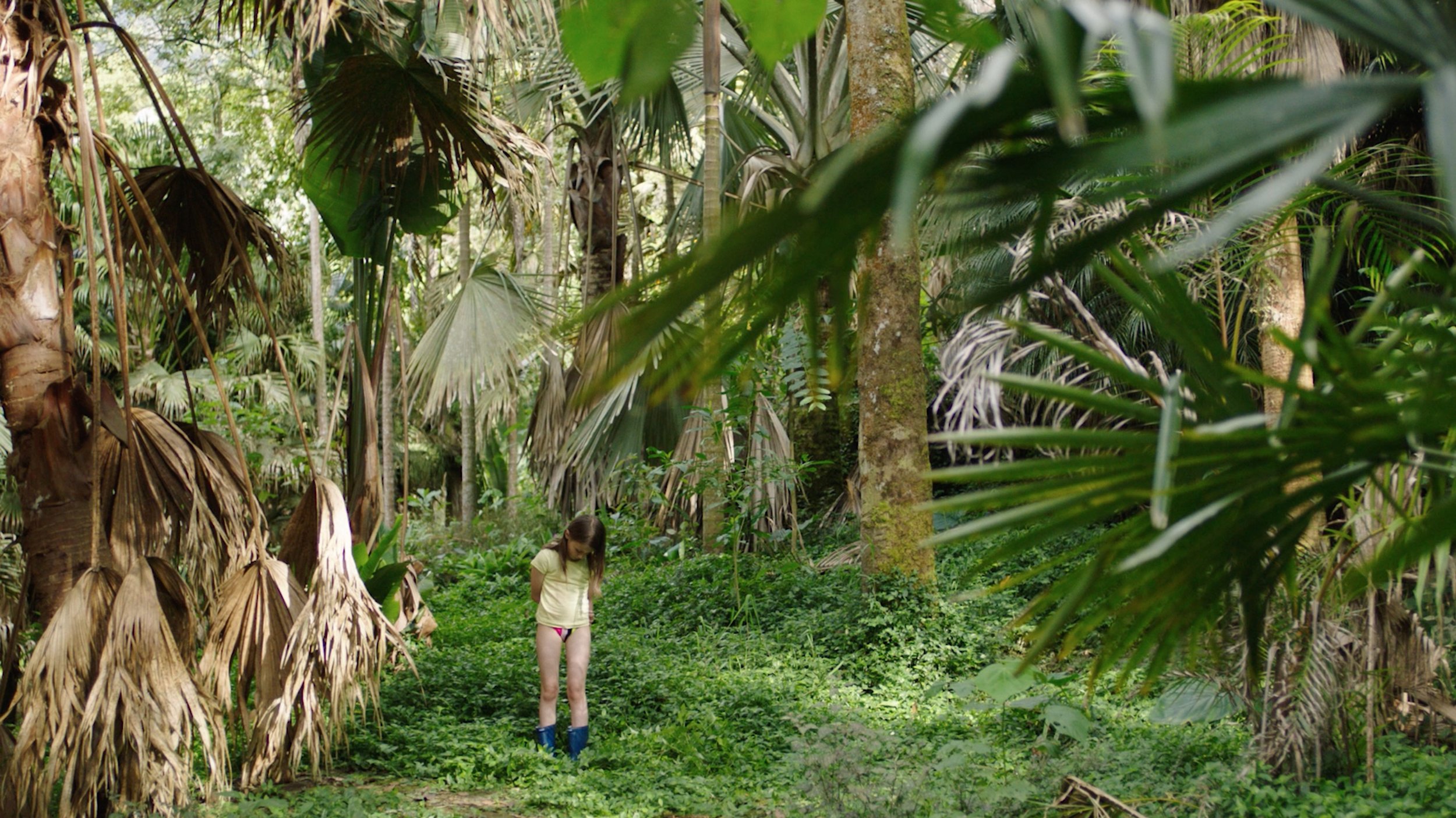 A little girl shown standing in rubber boots, underwear and a yellow t-shirt in the midst of a lot of plants and greenery outside during daytime. She looks lonely and isolated. 
