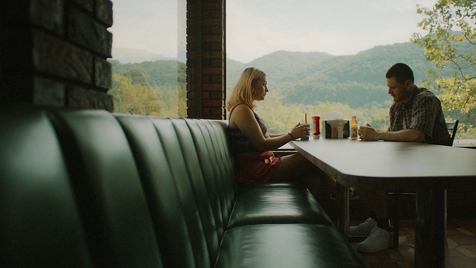 Nelly and Dane on their date. They are sitting in a corner of a room. Nelly is sitting on the end of a long dining couch whilst Dane is sitting on a regular stool. Nelly looks at Dane, while Dane is looking down at his hands resting on the table. Next to them is a large window which shows beautiful nature of many trees in the distance in green and yellow. It's less lonely and more intimate. 