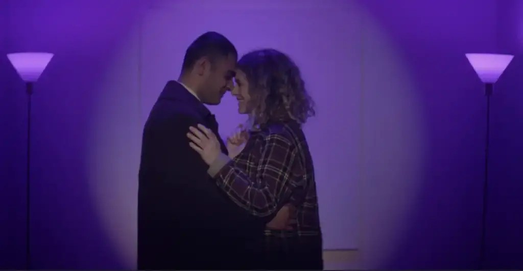Anabel (Anna Maguire) and George (Hamza Haq) embracing, surrounded by soft purple lighting.