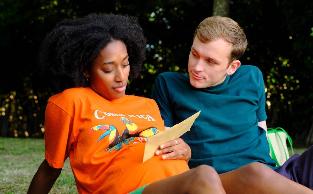 Ben (Harry Lawtey) and Jess (Sophia Brown) in the blissful before. They are sitting out in a park, Jess holding a letter with her hand whilst resting said hand on her pregnant belly. Ben is looking at her with a slight smile. The still is warm and colourful.  
