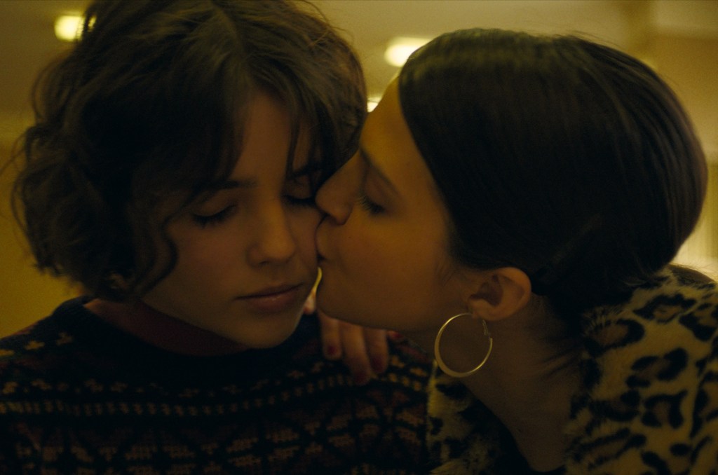 Close up of Tonia and Lera, as Tonia has her eyes closed and Lera kisses her on the cheek. Lera is wearing a coat with a leopard pattern and gold hoop earrings. Tonia is wearing a muted patterned jumper. 