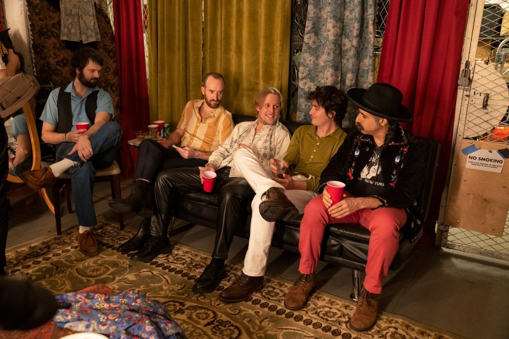 John Early and his four male band members sitting backstage drinking from red plastic cups. John is slouched back on the sofa, the one talking out of the group, as the others look towards him and listens. 