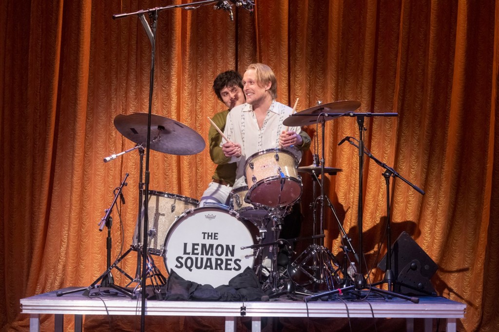 John Early sitting in the lap of Will Lawrence (the drummer) during rehearsal. Will is behind John, holding his hands that are holding the drum sticks. It's as if Will is teaching John to play the drums. Will is looking at John while John looks away and smiles happily.