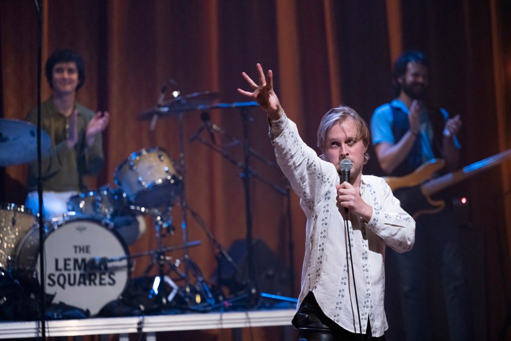 John Early on stage during his performance. His drummer and bass player is seen in the background out of focus applauding him. John is holding his microphone with his left hand close to his mouth while his right hand is outstretched towards the balcony (not shown). He is looking up towards the same direction.