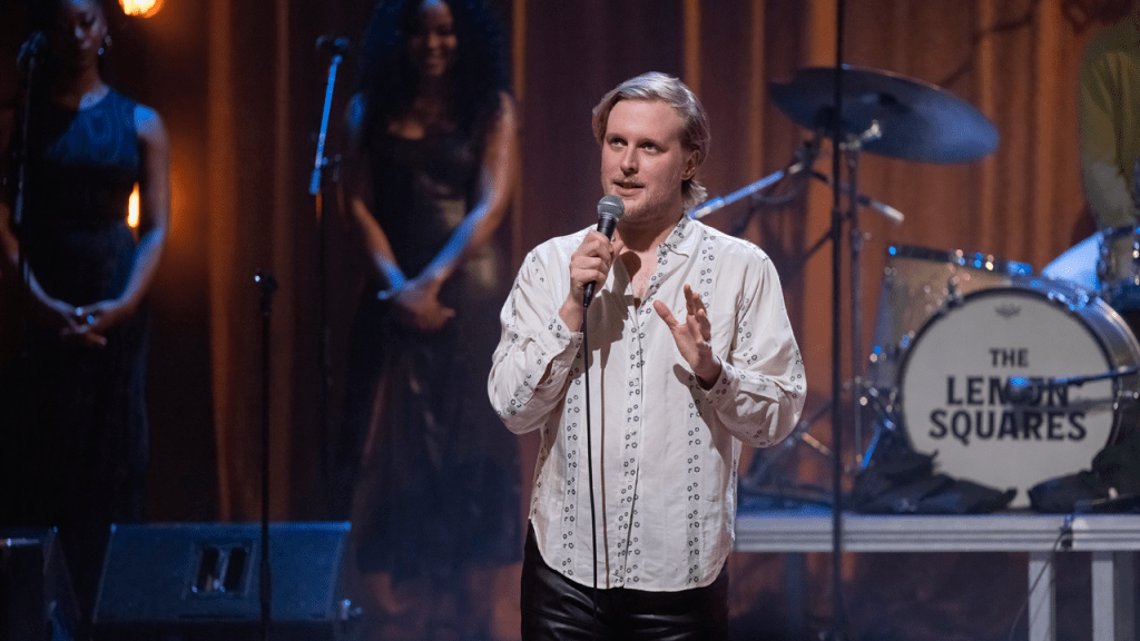 John Early standing on stage in the midst of talking, with the microphone close to his mouth. One hand is holding the microphone, the other is doing a gesture. In the background his two backup singers are seen looking towards him at the still's left and the drum head saying "The Lemon Squares" is seen to the still's right.
