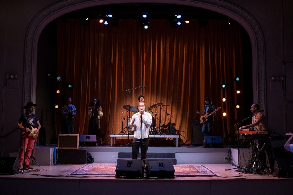 John Early and his band seen rehearsing on stage. Still taken from afar, the whole stage is visible within the frame as are all band members. There's a spotlight making Early stand out as the other band members are positioned surrounding him. 
