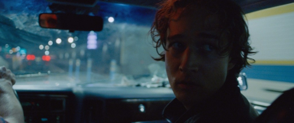 Still taken from the backseat of a car, showing Jack sitting in the passenger's seat, turning his head back as if looking at someone following them by car. He has a serious facial expression. In front of the car, blinking lights are seen and it's evident that they are on the move.