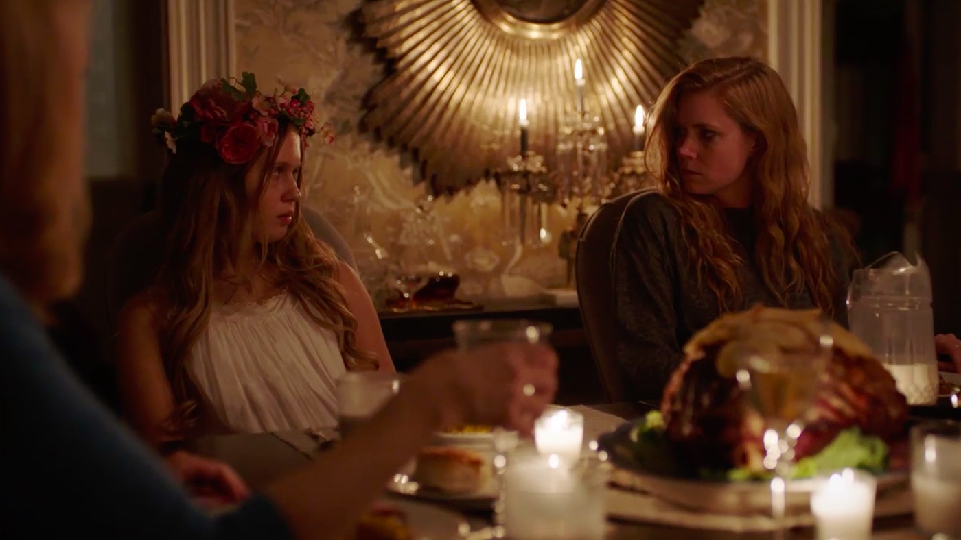 A still from the miniseries 'Sharp Objects' that shows Amma (Eliza Scanlen) and Camille (Amy Adams) sitting next to one another at an ornate dinner table. Amma wears a flower crown and a white dress while Camille wears a
