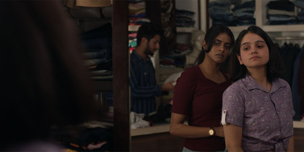 In a clothing store, Anila and Mira are seen standing in front of a mirror, both inspecting Mira's lilac patterned shirt with short sleeves. They both seem to be very focused in their facial expressions and their eyes are focused on the reflection in the mirror. In the same mirror, a male worker can be seen out of focus whilst handling a garment. 