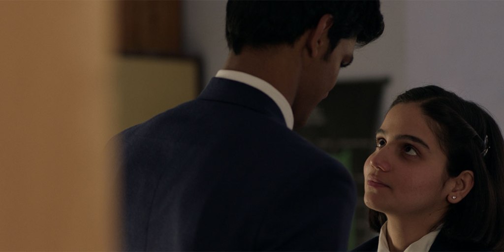 Close-up showing Mira's face as she is looking up towards the taller Sri that is standing in front of her with his back turned towards the camera. She looks at him with curiosity but also with a bit of innocence. Since the camera is also slightly more aligned with Mira's height, the emphasis of Sri's length (as well as experience/maturity) is further underlined in this specific framing as he is looking down at Mira. 