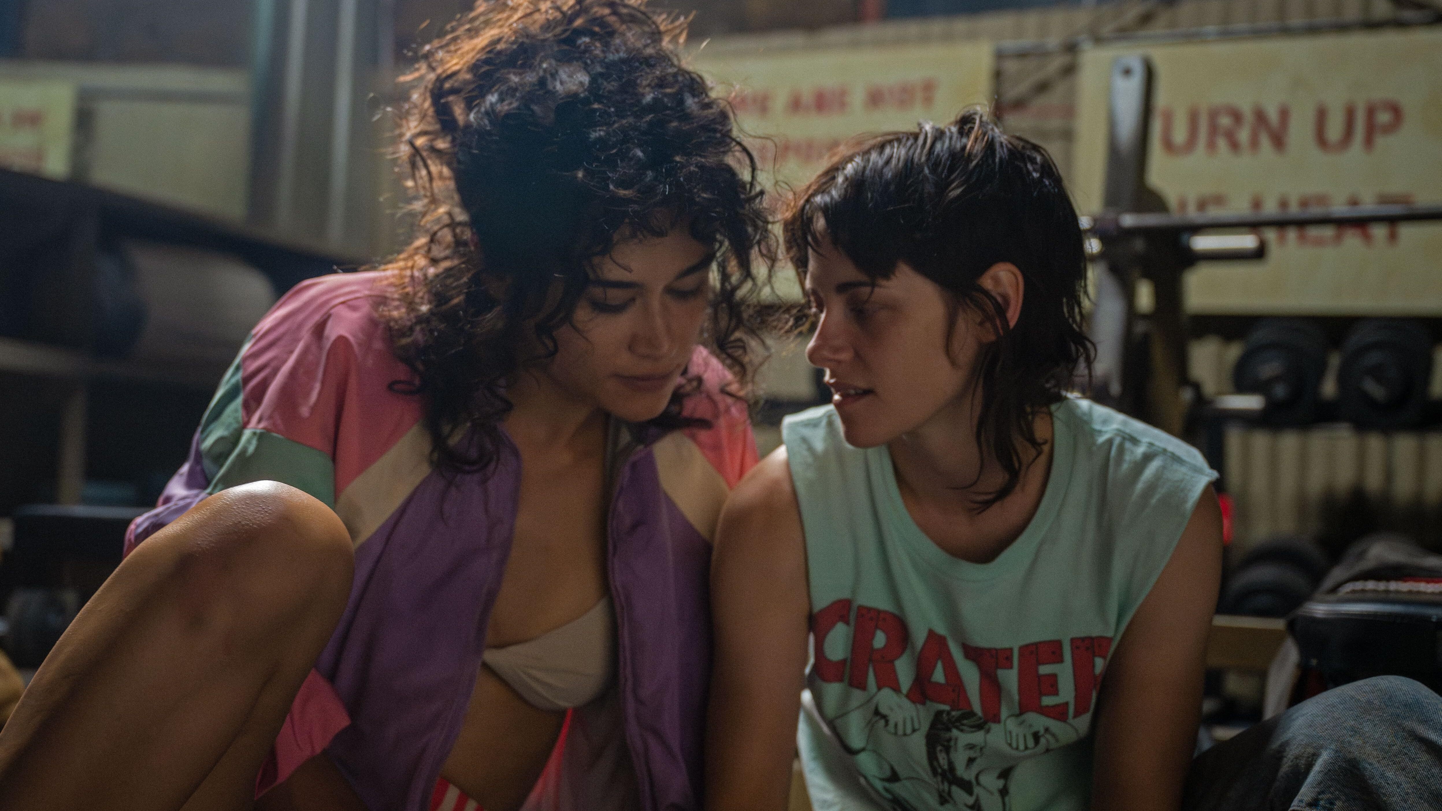 A still of Jackie (Katy O'Brian) and Lou (Kristen Stewart) in 'Love Lies Bleeding'. Jackie wears a colourful bomber while Lou wears a muscle tank. The two are in the weight room of a gym.