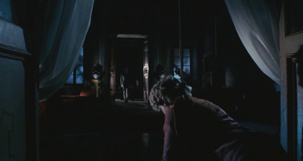 Still from A Bay of Blood. Interior shot at nighttime with no visible source of light. In the foreground, an older woman with a nose around her neck is seen from behind. In the distance at a doorway, a man is seen hidden in the shadows. 