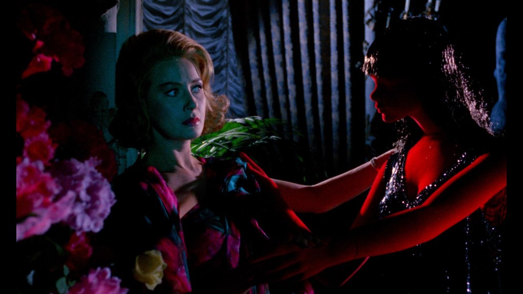 Still from Blood and Black Lace. A woman is seen looking frightened at a red mannequin. The still bathes in red, lilac and green hues. 
