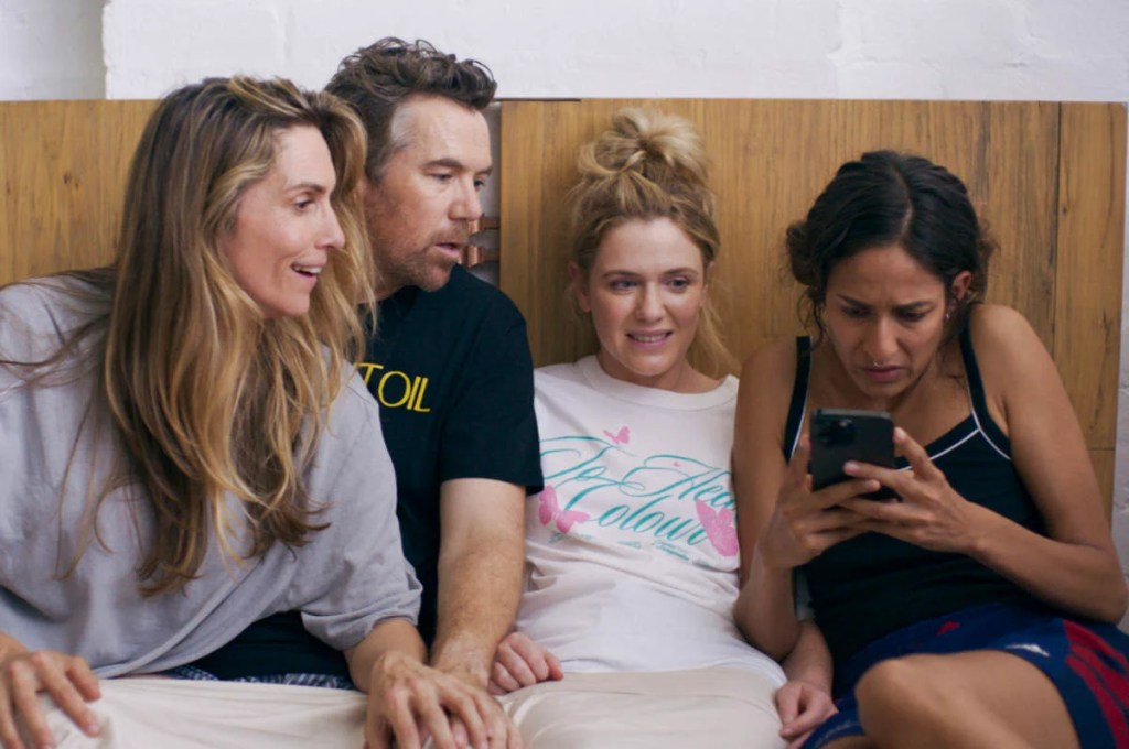 Still from Colin from Accounts. In order of appearance: Chiara, Gordon, Ashley and Megan are in bed. All are looking towards Megan who is reading something on her phone with a concerned facial expression. Chiara looks happy, Gordon looks curious and Ashley looks uncertain with a more ambiguous smile.