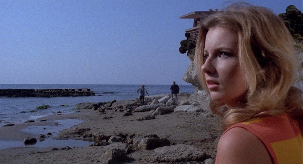 Still from Five Dolls for an August Moon. A young woman is seen in the foreground to the right. Behind her in the distance the outline of a house is seen as well as two men walking across the sandy and stony beach. 