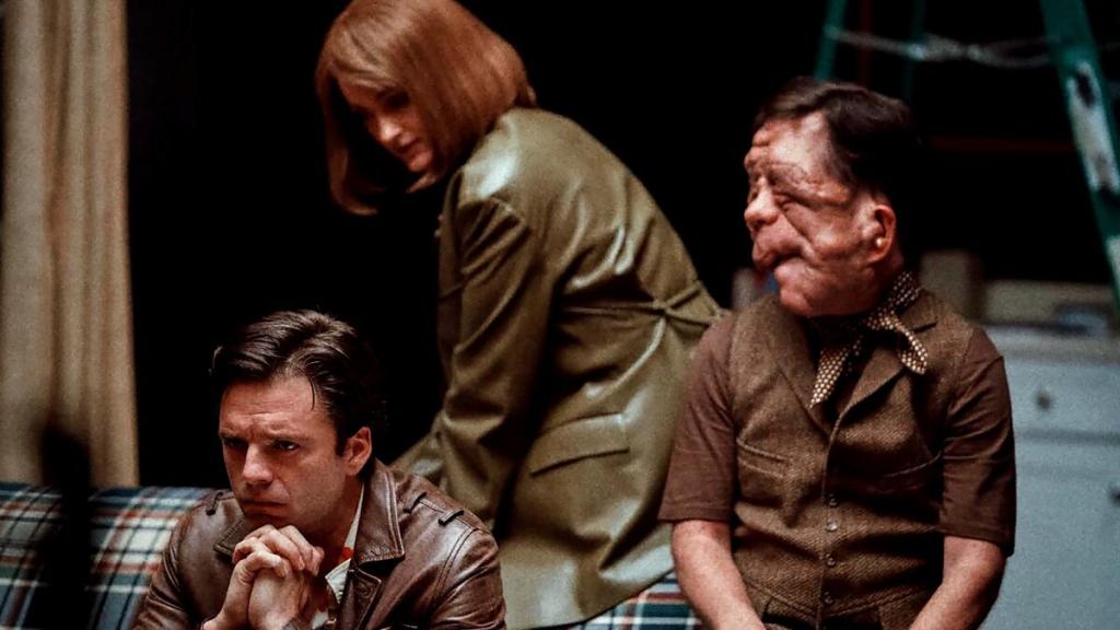 Sebastian Stan, Renate Reinsve, and Adam Pearson in a still from the film 'A Different Man'. Stan and Pearson wear similar brown attire, with Reinsve in a green leather jacket between them.