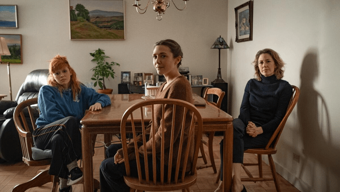 A still of Natasha Lyonne, Elizabeth Olsen, and Carrie Coon in 'His Three Daughters'. The three women sit on chairs in a kitchen, facing the camera.
