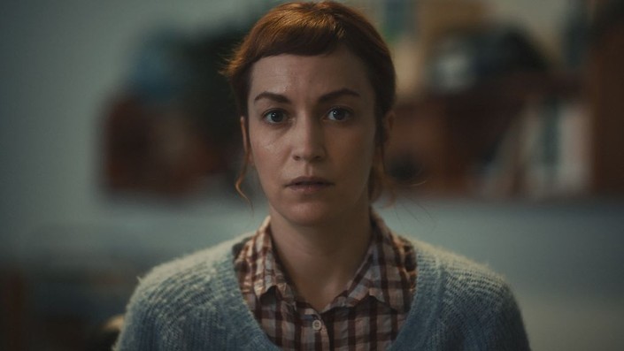 A still of Britt Lower as "Miriam Gordon" in the film 'Darkest Miriam'. She is in focus as she stands in the centre of the frame and wears a checkered flannel with a sweater over top.