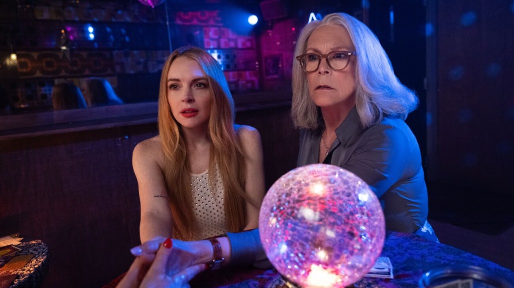 A still of Lindsay Lohan and Jamie Lee Curtis in 'Freakier Friday'. The two women are in the centre of the frame, with purple lighting around them and a crystal ball between them.