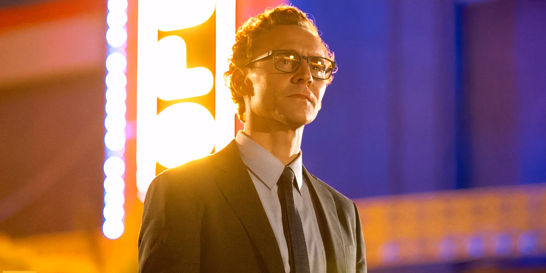 A still of Tom Hiddleston as the title character in the film 'The Life of Chuck'. He wears glasses and a suit, and stands in focus in front of a brightly lit marquee.