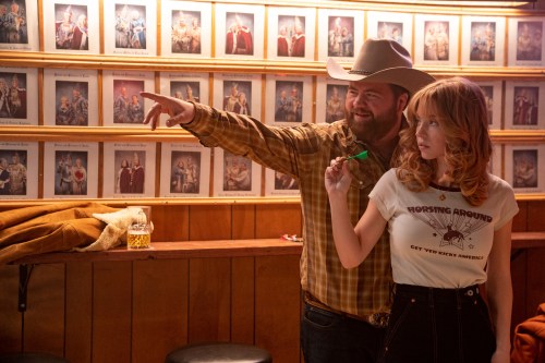 A still of Lefty (Paul Walter Hauser) and Penny Jo (Sydney Sweeney) playing darts at a bar. Lefty is wearing a cowboy hat and plaid shirt, and gestures with his right hand. Penny Jo is wearing a white t-shirt that reads "Horsing Around: Get Yer Kicks America" and high-waisted jeans, looking to the side as she holds a green dart.